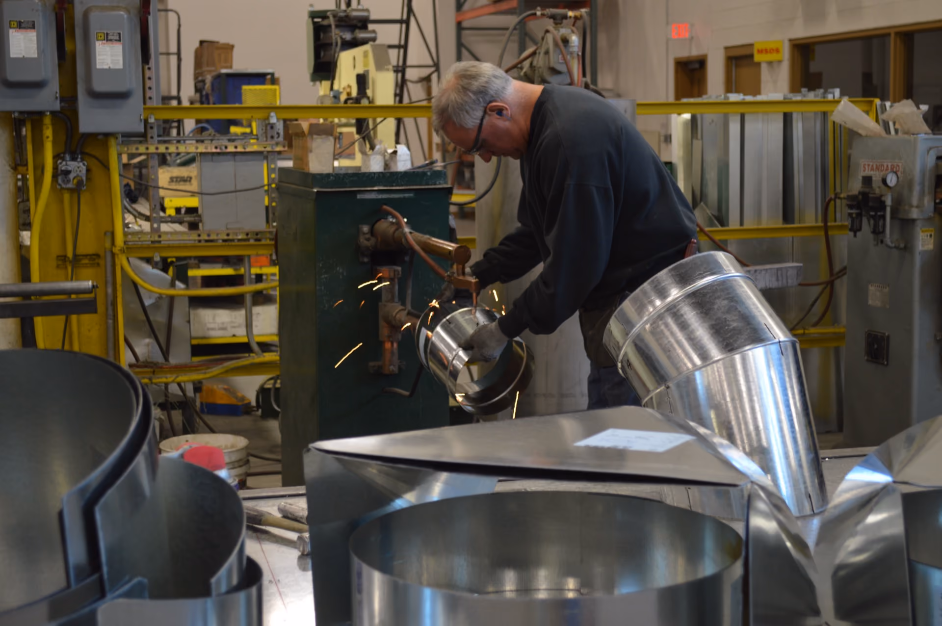 Man using welding equipment to work on shiny metal duct pieces in an industrial workshop.