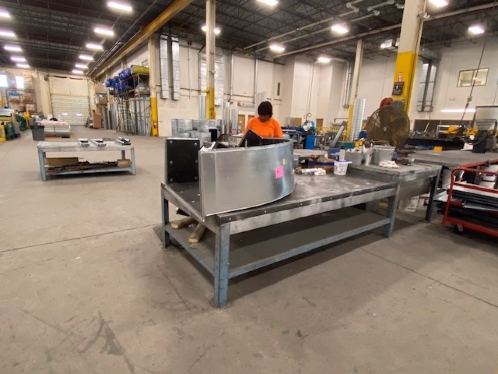 Worker in orange shirt inspecting metal duct component on a large industrial workbench inside a factory.