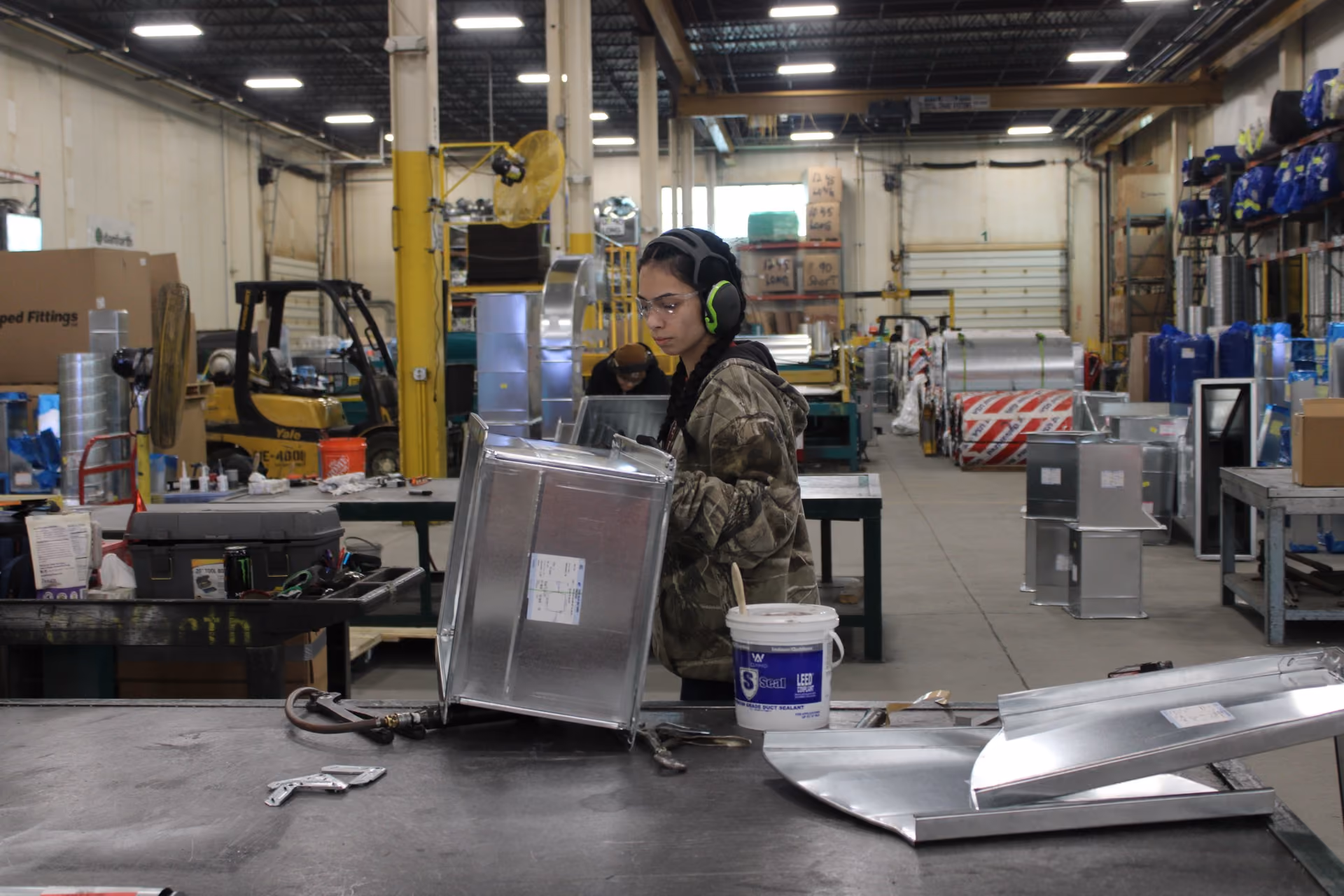 Worker wearing earmuffs and safety glasses handling metal duct pieces in a large industrial workshop.