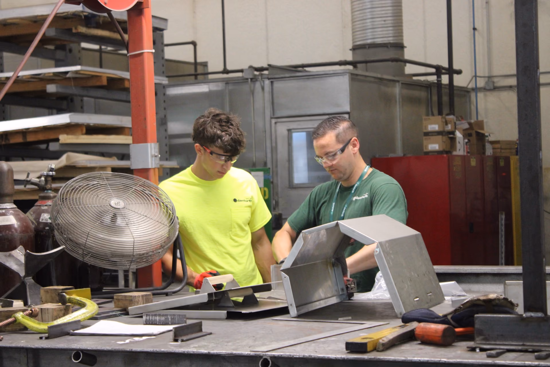Two men in safety glasses working on metal parts at a workshop table with tools around.