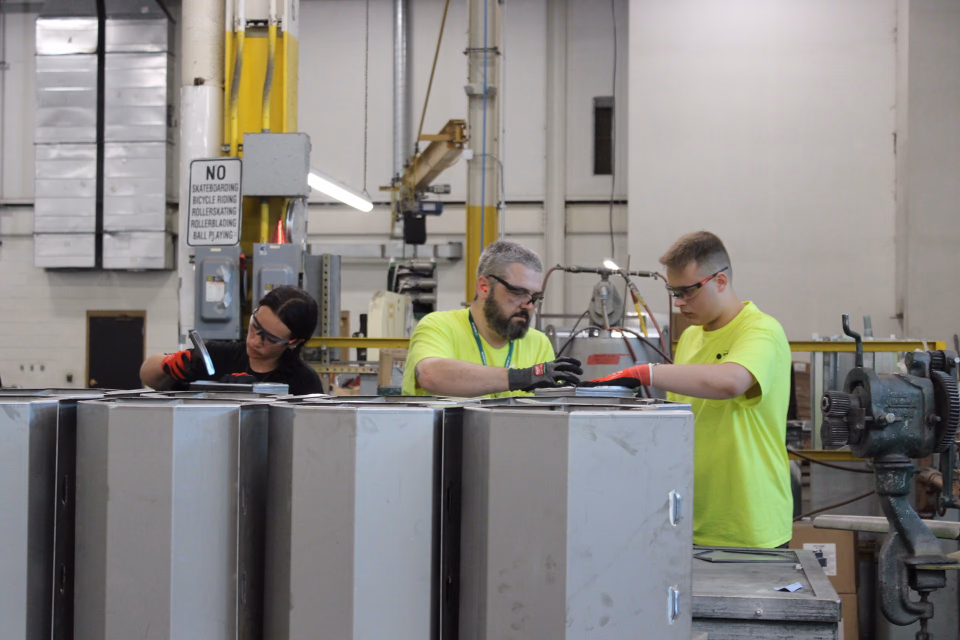 Three factory workers wearing safety glasses and gloves assembling metal parts indoors.