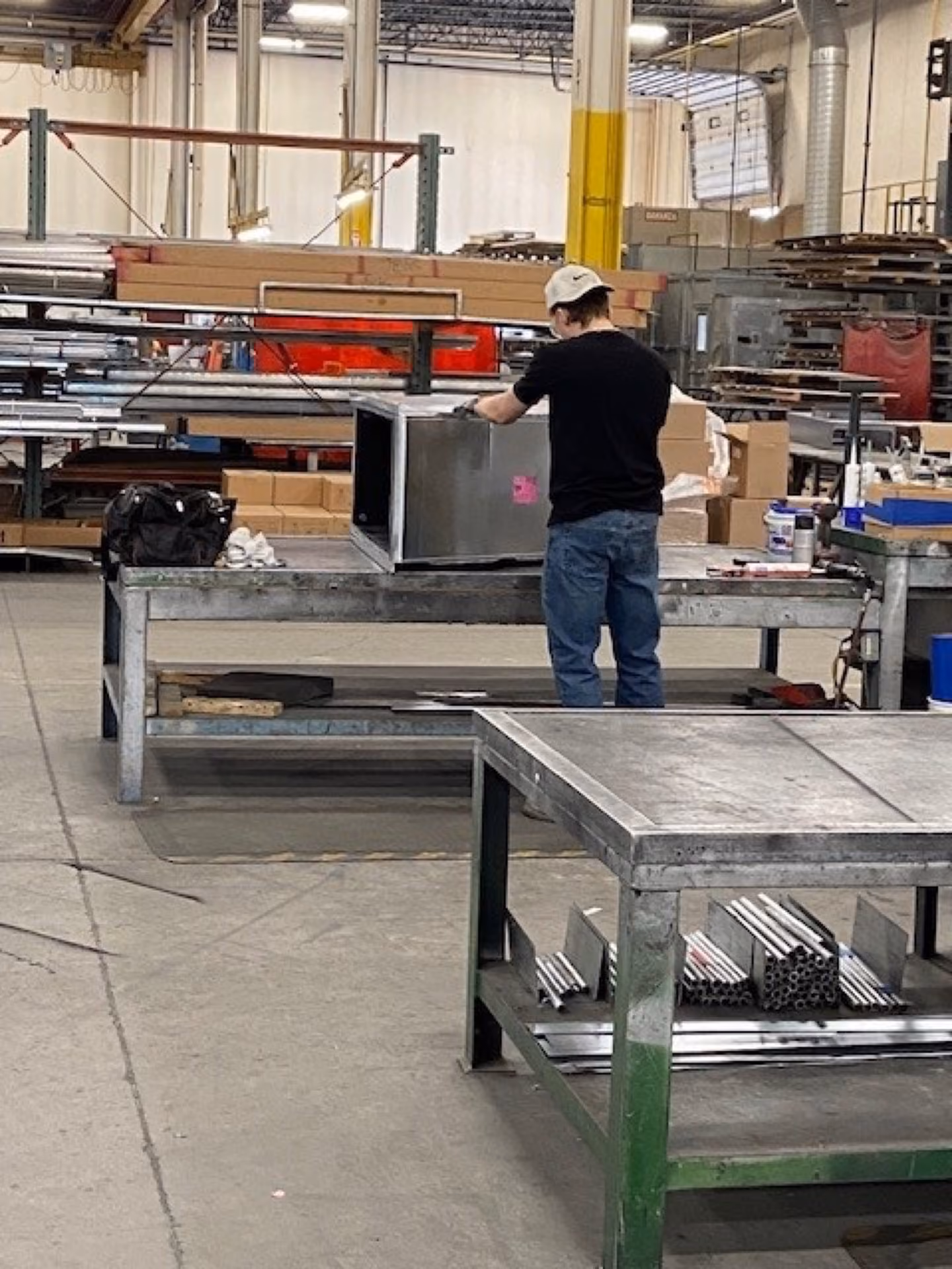 Man in a workshop wearing a cap and gloves working on a metal box on a steel table.
