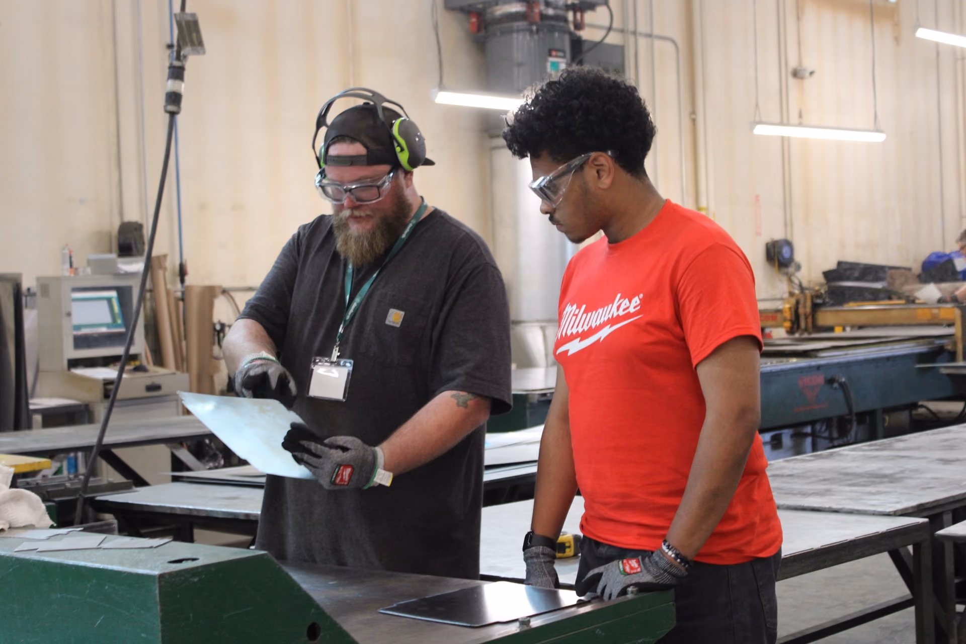 Two men in safety glasses and gloves examine metal sheets in a workshop with industrial equipment.