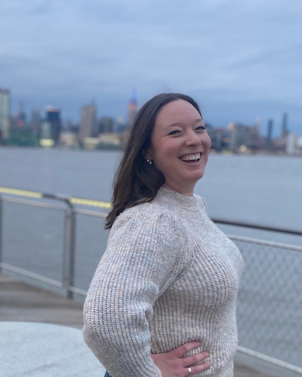 Kelly, smiling, on the waterfront with the Manhattan skyline behind her
