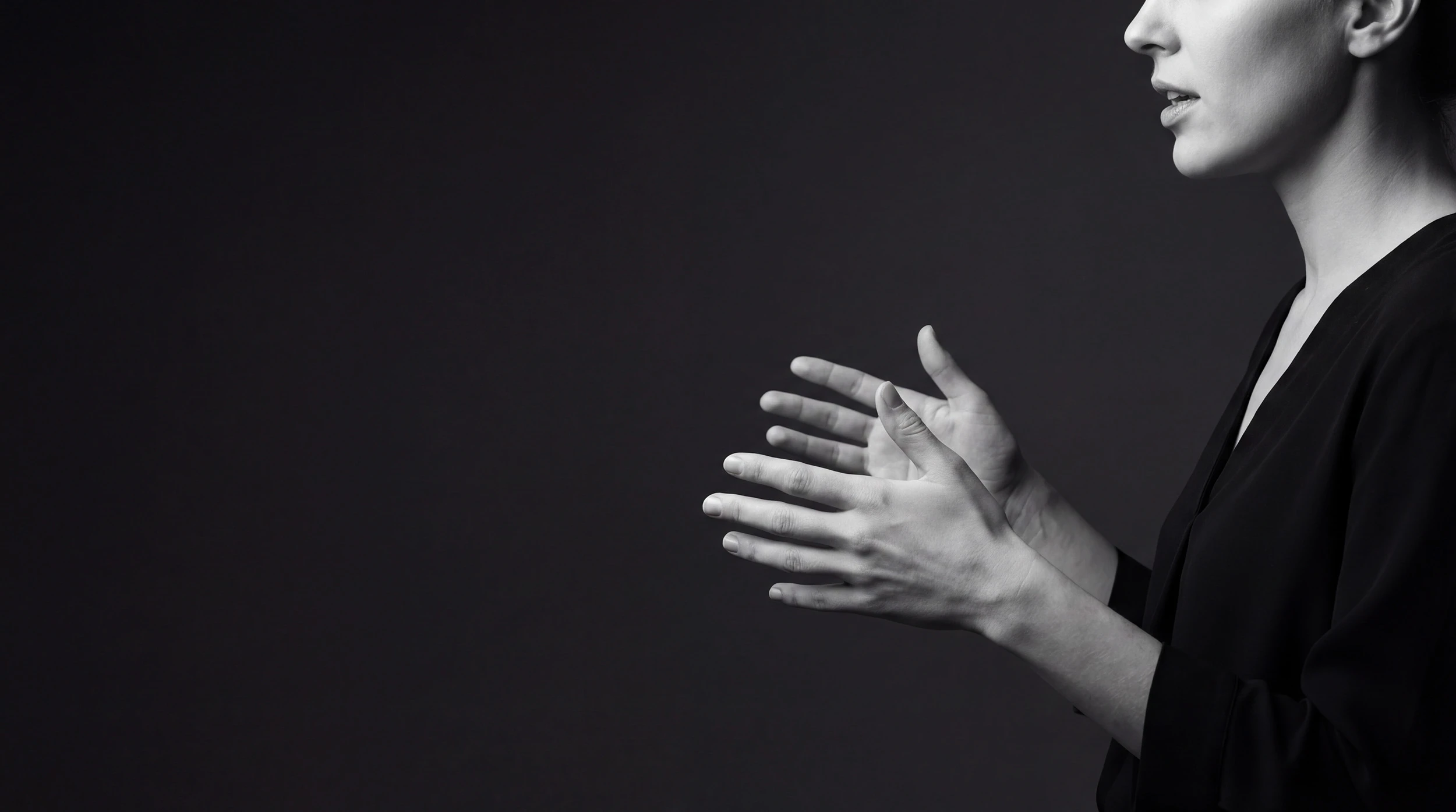 Side view of a woman in black clothing gesturing with her hands against a dark background.