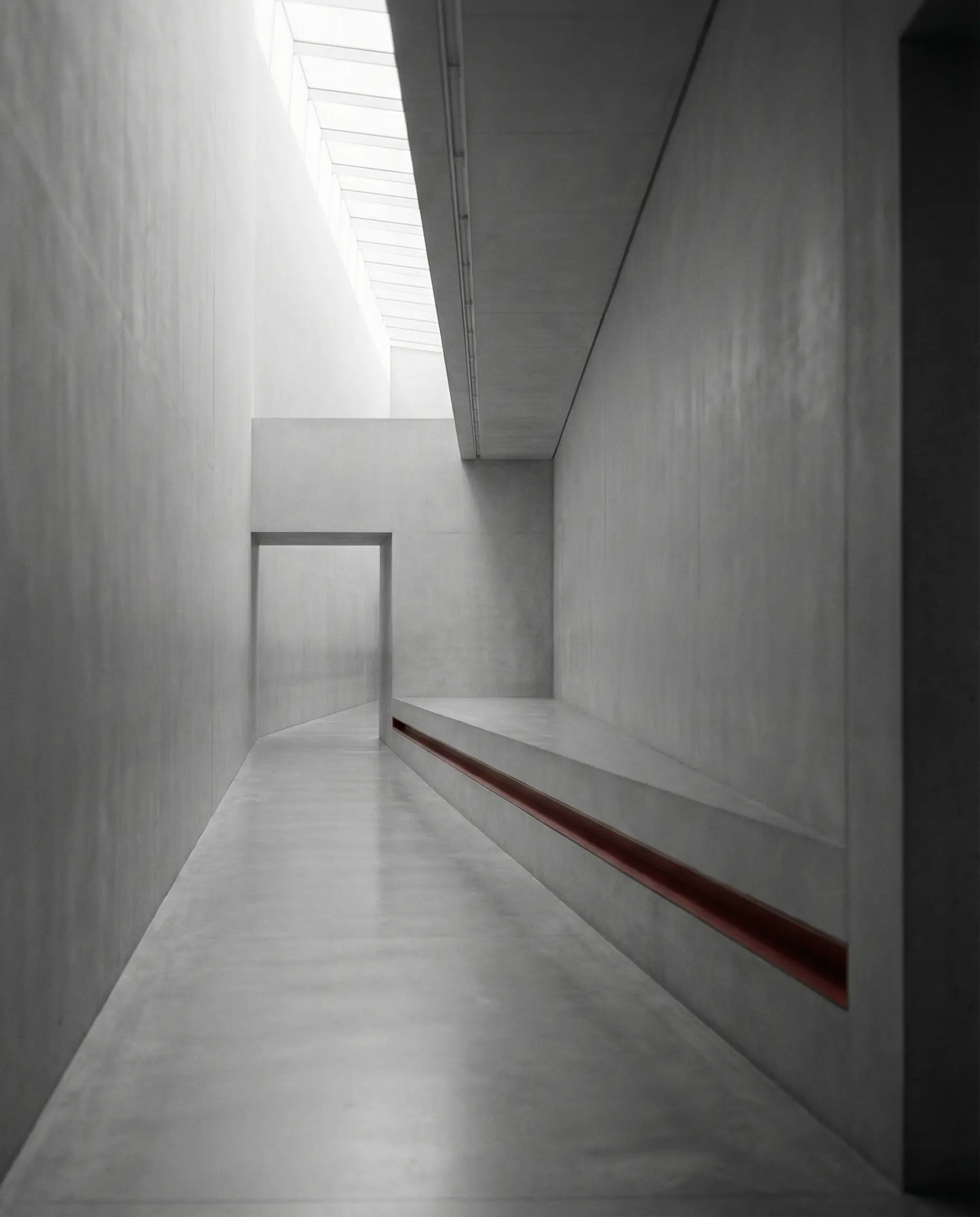 Minimalist concrete interior corridor with skylight, angled bench, and rectangular doorway.
