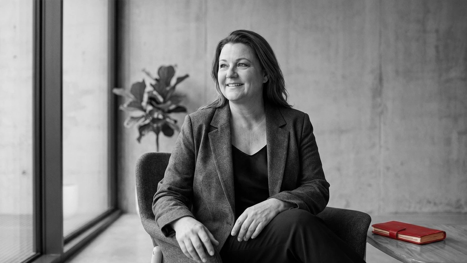 A woman with shoulder-length hair sitting in a chair by a large window, smiling and looking to the side, wearing a blazer over a dark top, with a red notebook on a nearby table.