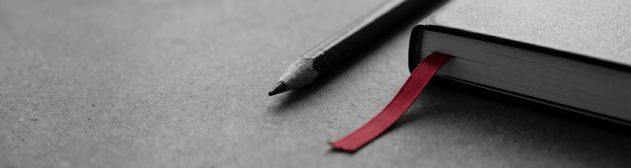 Close-up of a pencil next to a closed book with a red ribbon bookmark on a textured surface.