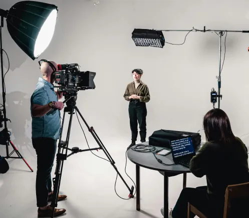 Person stands in a studio being filmed by a cameraman, with a director reviewing a script on a laptop.