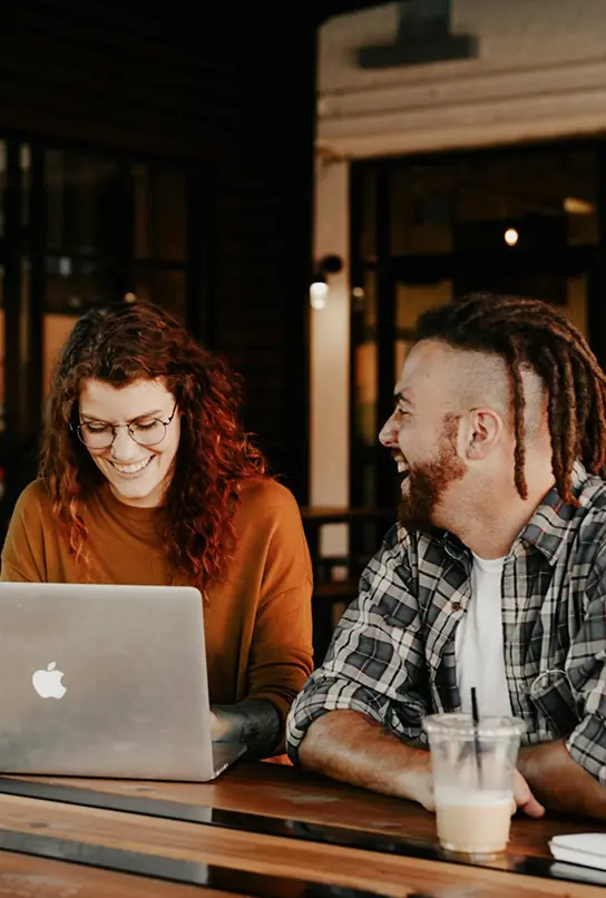 Two people smiling and chatting at a table with a laptop and iced coffee in a cozy setting.