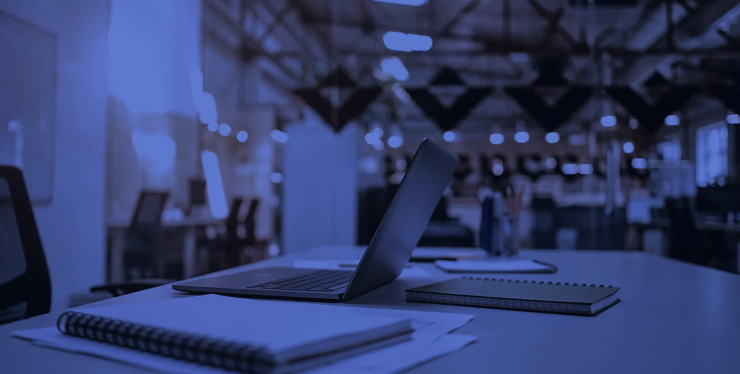 Modern office workspace with an open laptop, notebooks, and stationery on a long white table in a bright, industrial-style office environment.