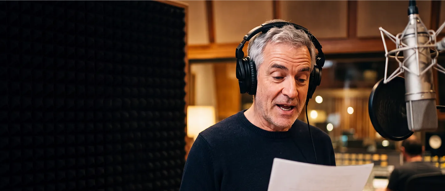 A middle-aged man with gray hair wearing headphones is reading a paper while speaking into a large professional microphone in a recording studio.