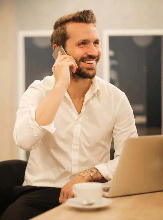 Smiling man with beard and tattoos sitting at a desk, talking on a smartphone with a laptop and coffee cup in front of him.