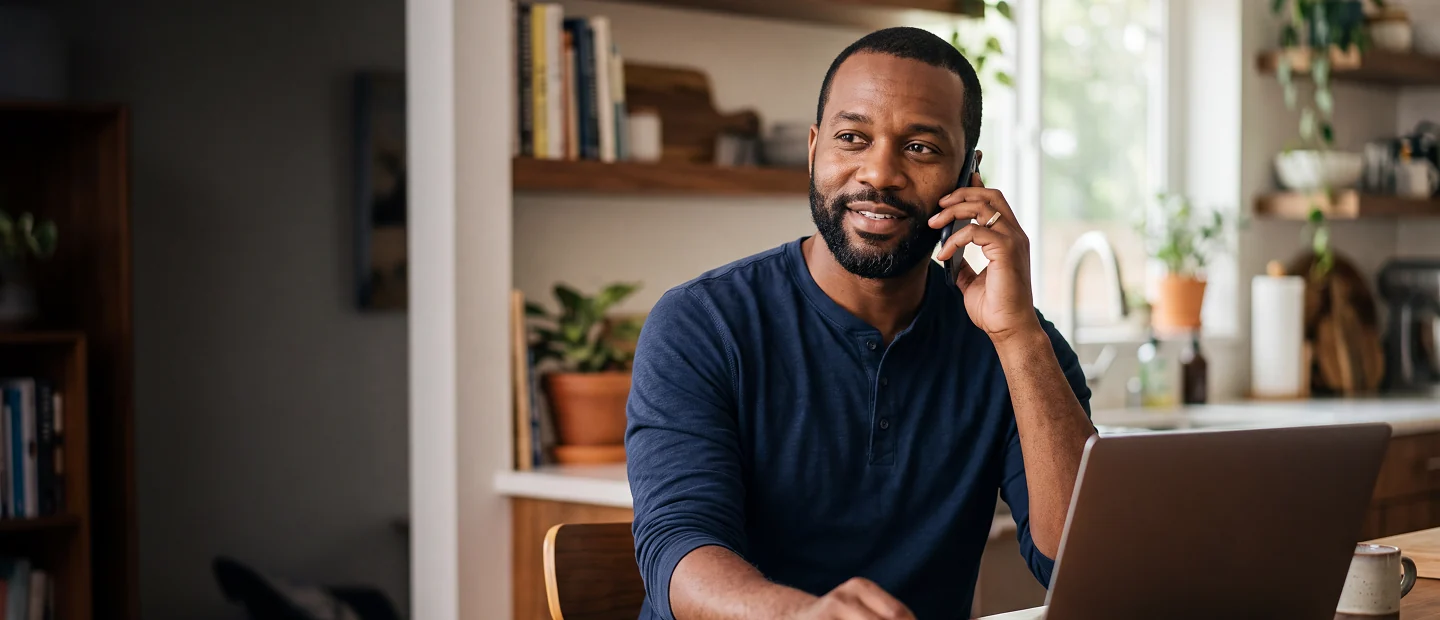 Smiling man wearing a navy blue long-sleeve shirt talking on a smartphone while sitting at a kitchen table with a laptop in front of him and plants and kitchen items in the background.