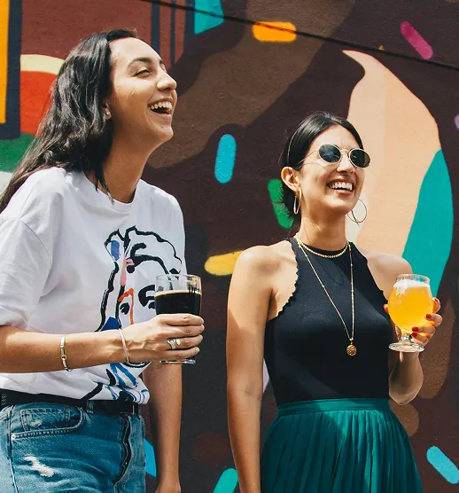 Two women laughing and holding drinks in front of a colorful mural outdoors.