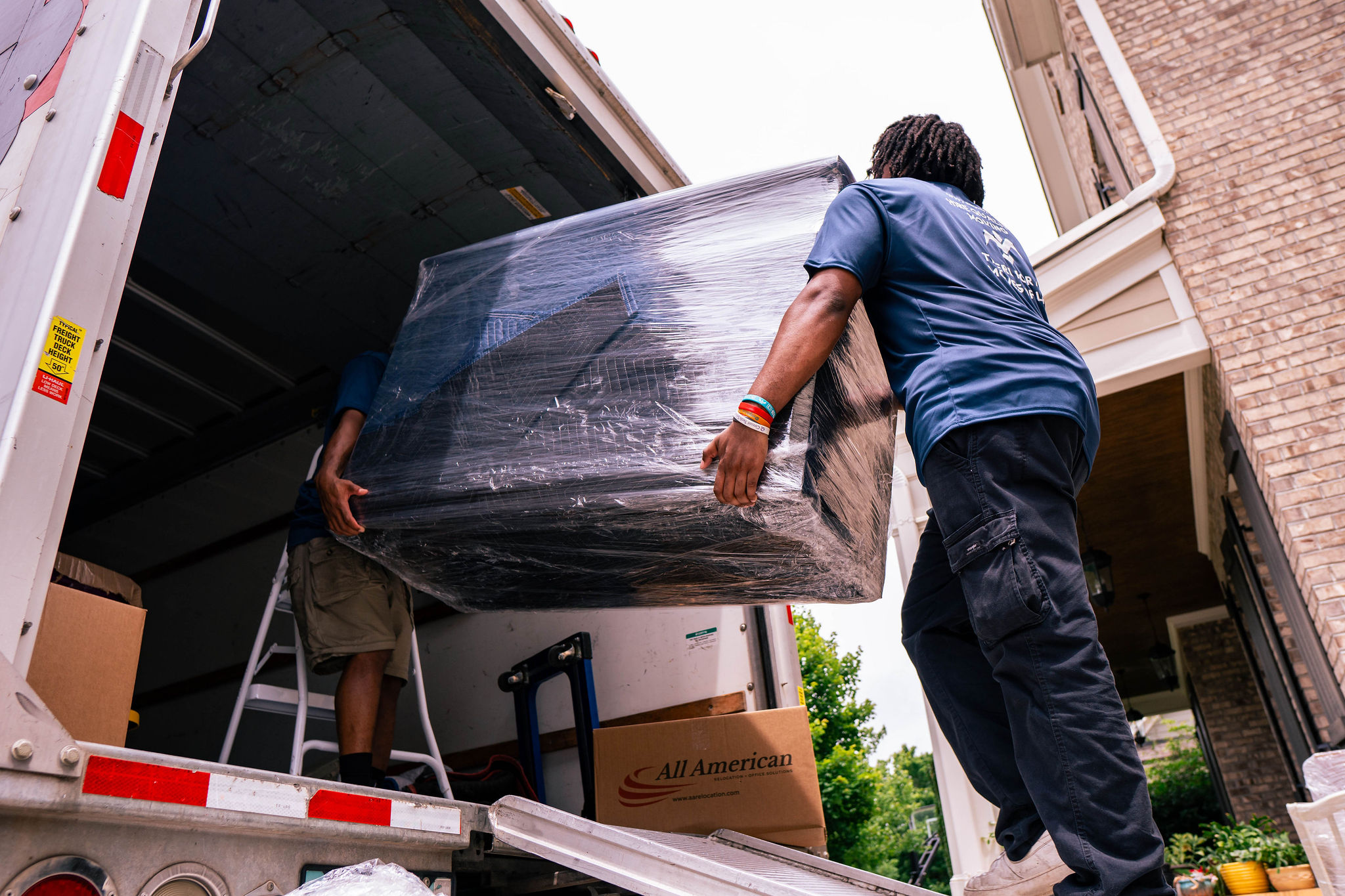 Two movers lifting a large wrapped furniture piece into a moving truck at a house driveway.