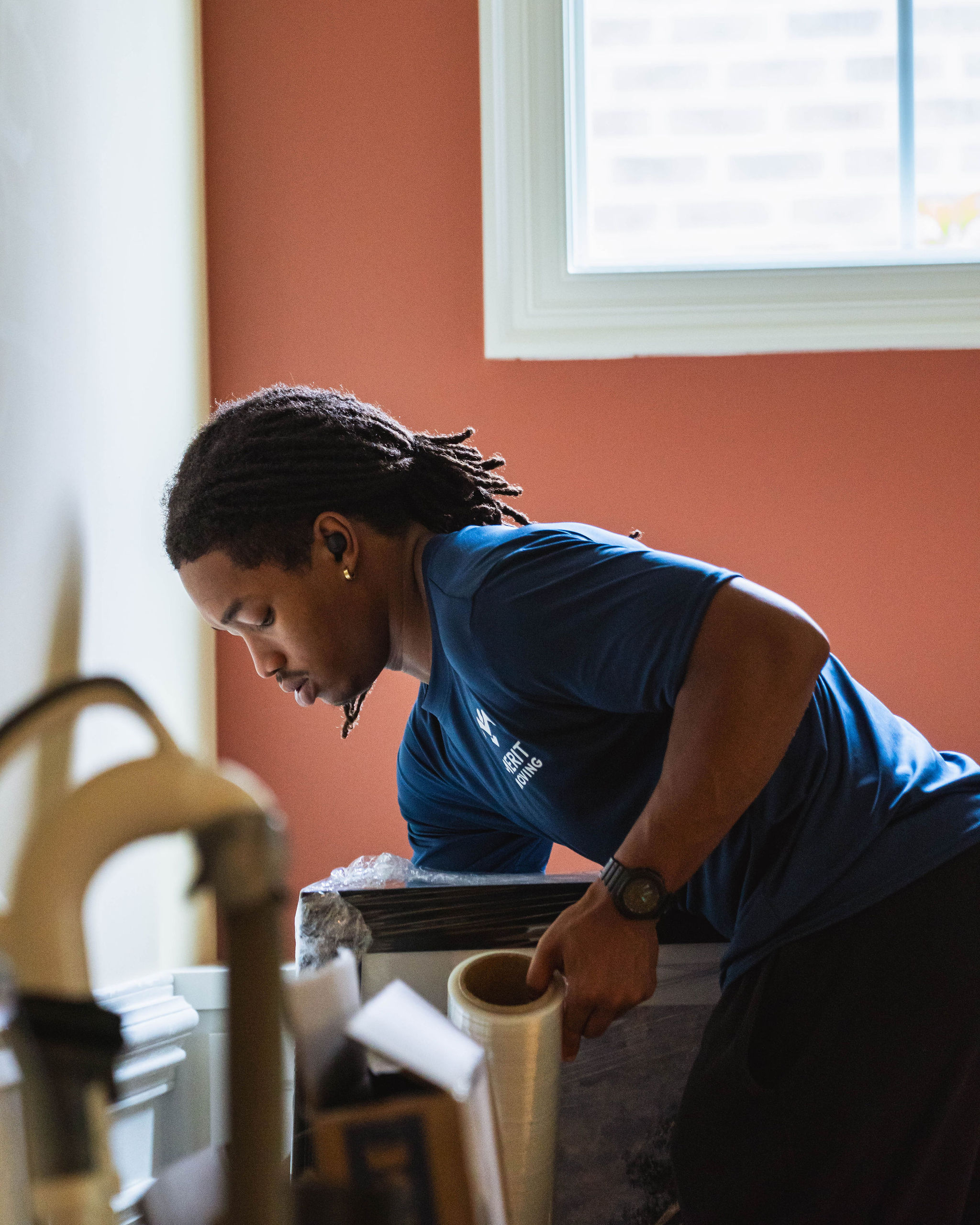 Man in a blue shirt packing items with plastic wrap near a window indoors.