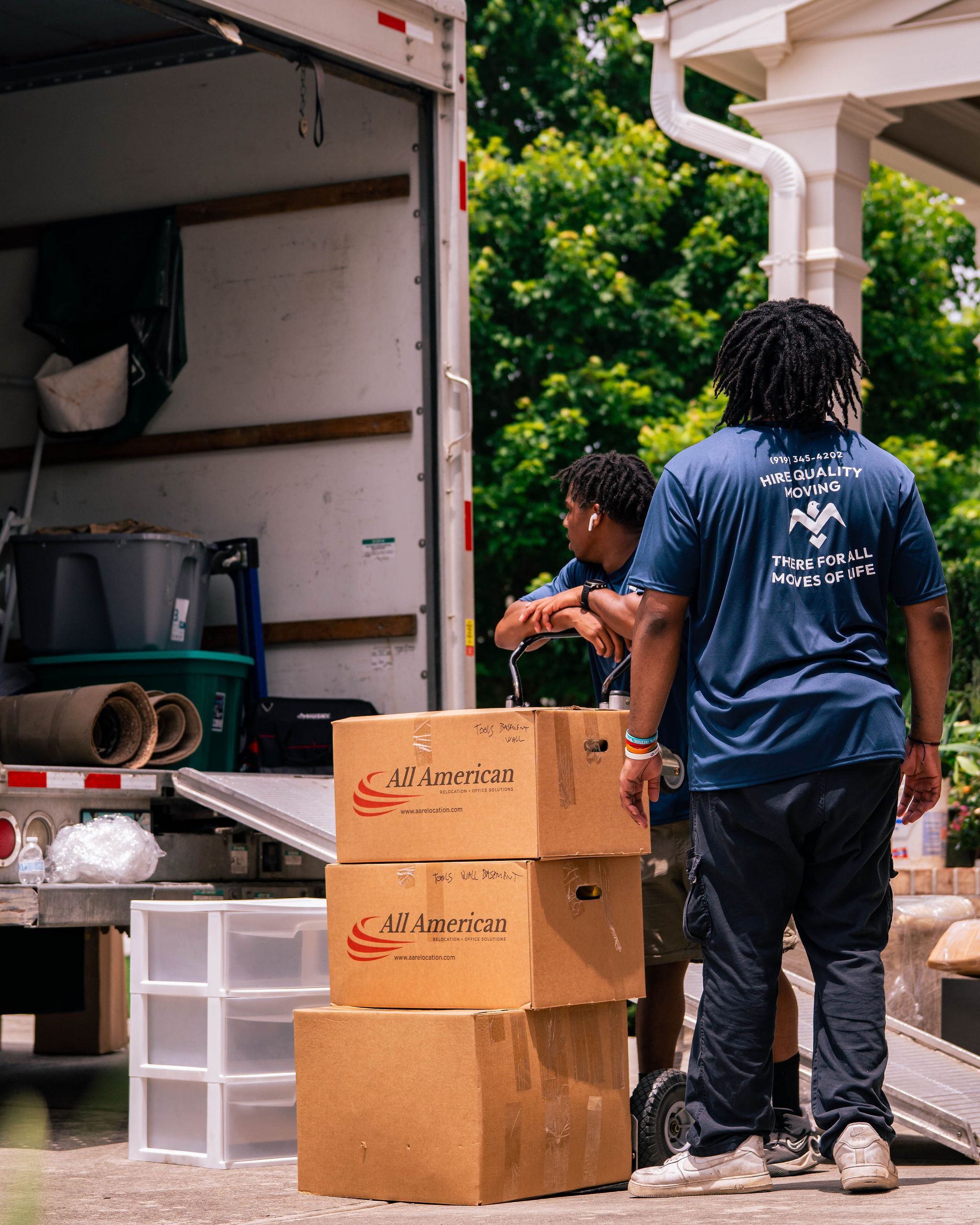 Two movers with a dolly near stacked cardboard boxes labeled All American next to an open moving truck.