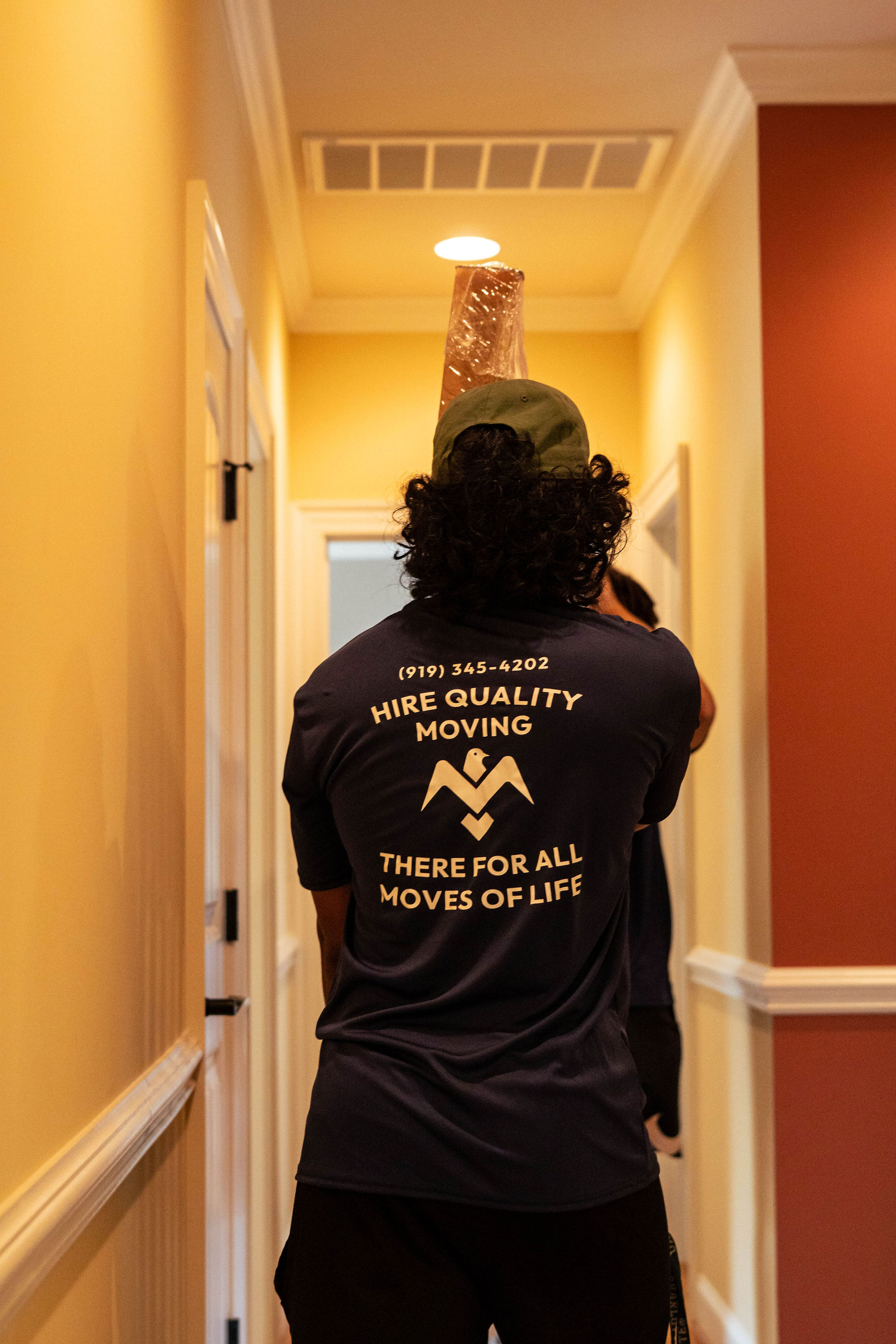 Mover wearing a 'Hire Quality Moving' shirt carrying an item wrapped in plastic through a hallway.