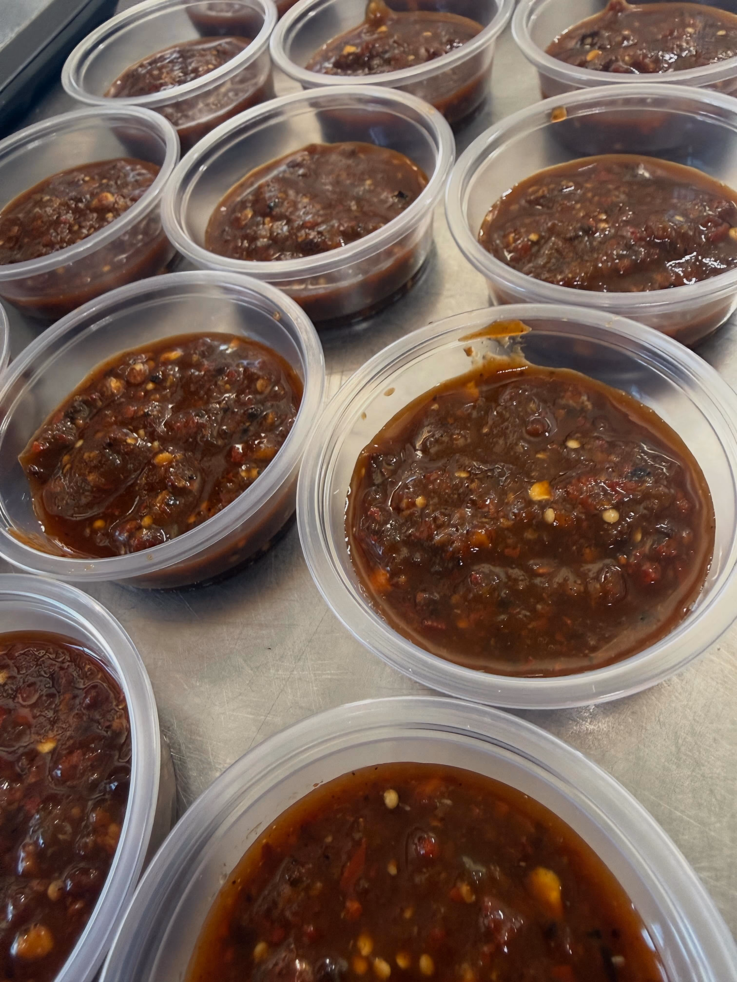 Plastic containers filled with dark red homemade chili sauce with visible chili seeds on a metal surface.