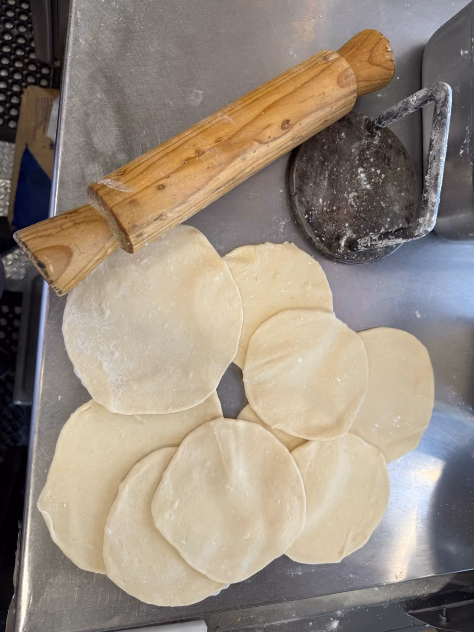 Wooden rolling pin and metal dough press on a stainless steel surface with several round pieces of rolled dough.