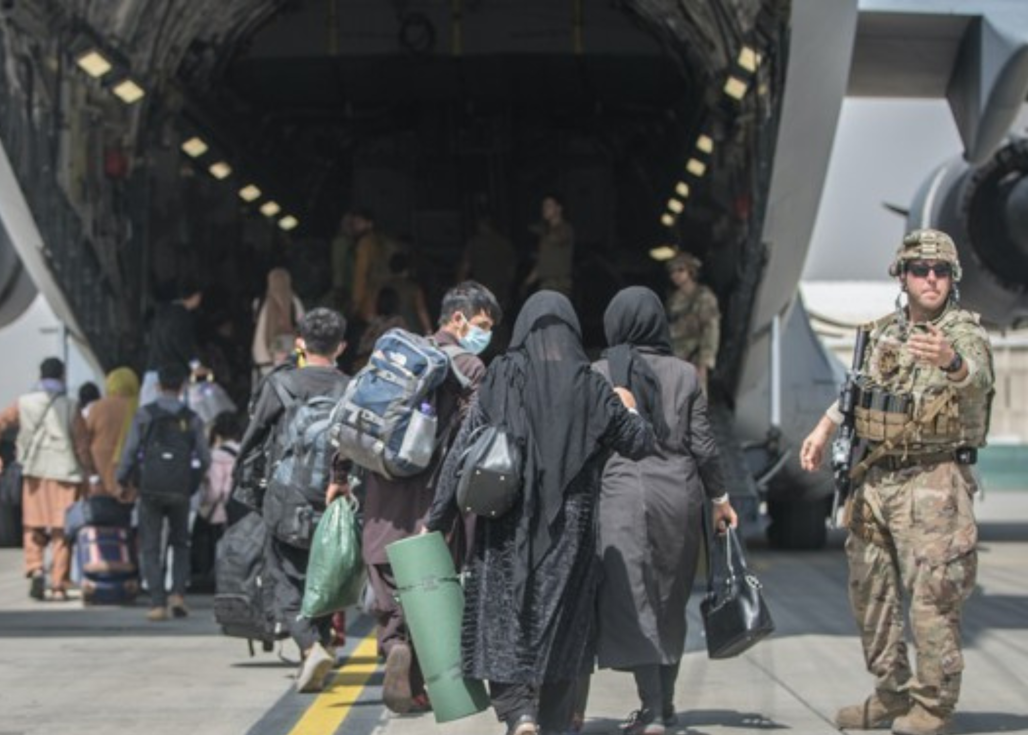 Families carrying luggage board a U.S. military cargo plane as a U.S. service member stands guard.