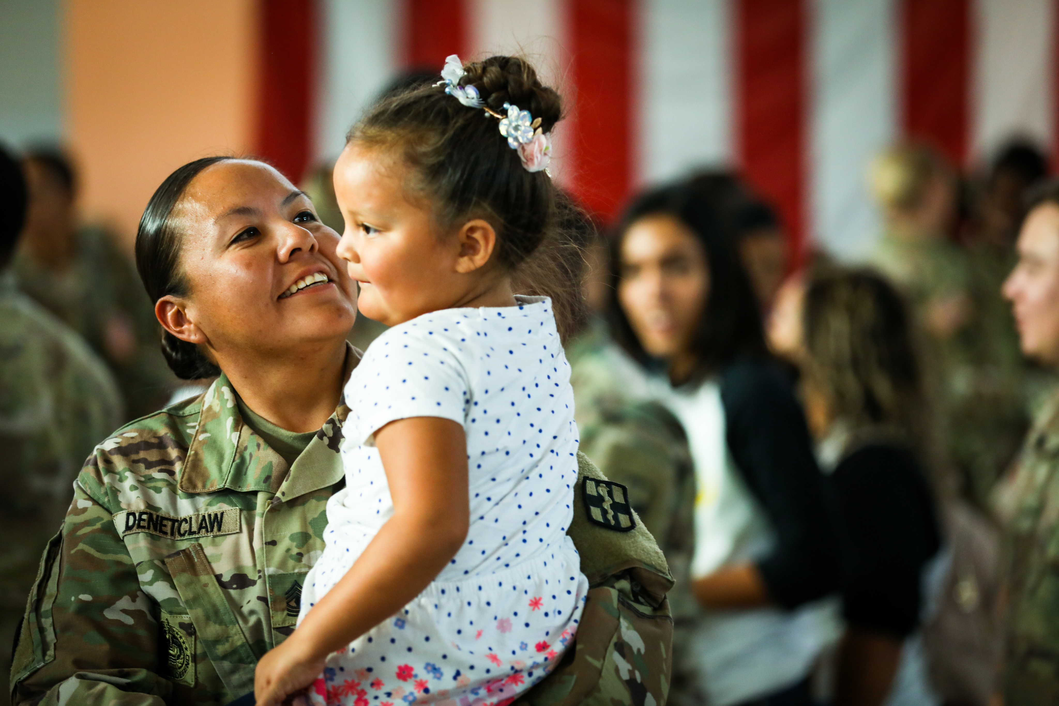 A U.S. soldier holds a young girl in front of a crowd and a large American flag.