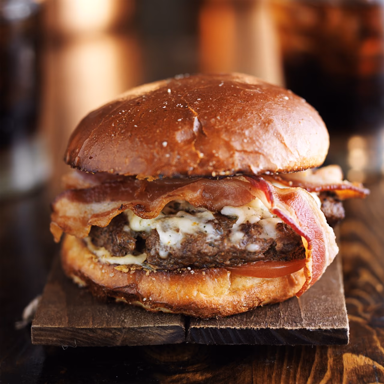 Bacon cheeseburger with melted cheese and tomato slice in a toasted bun on a wooden board.