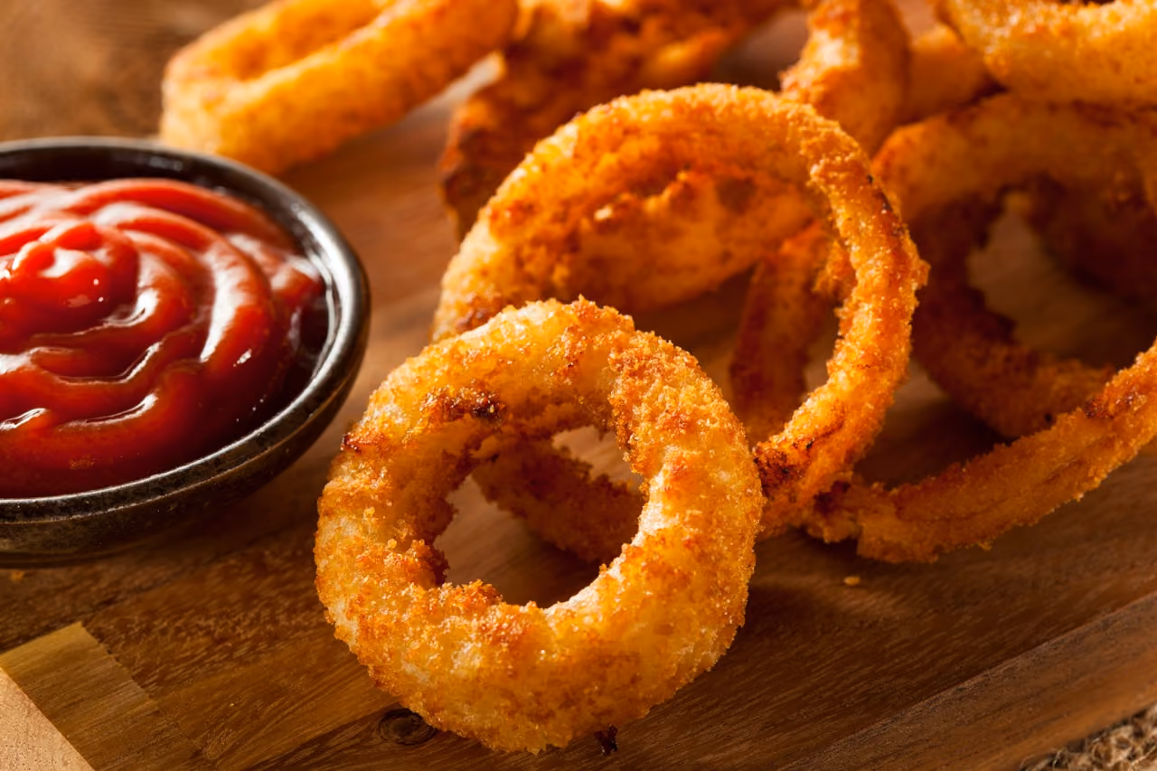 Close-up of crispy golden fried onion rings on a wooden board with a bowl of ketchup.