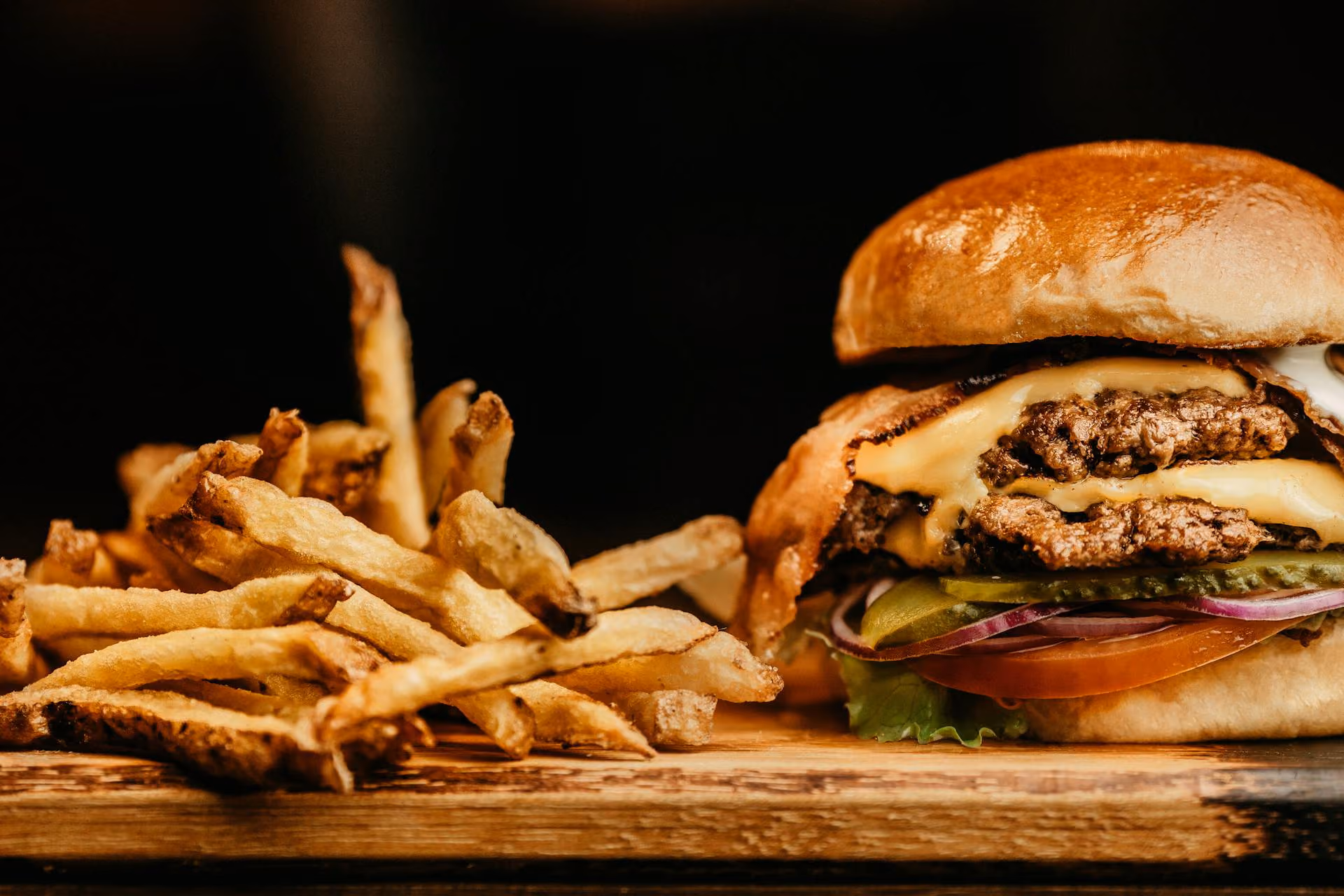 Cheeseburger with lettuce, pickles, tomato, onion, and fries on wooden surface with black background.