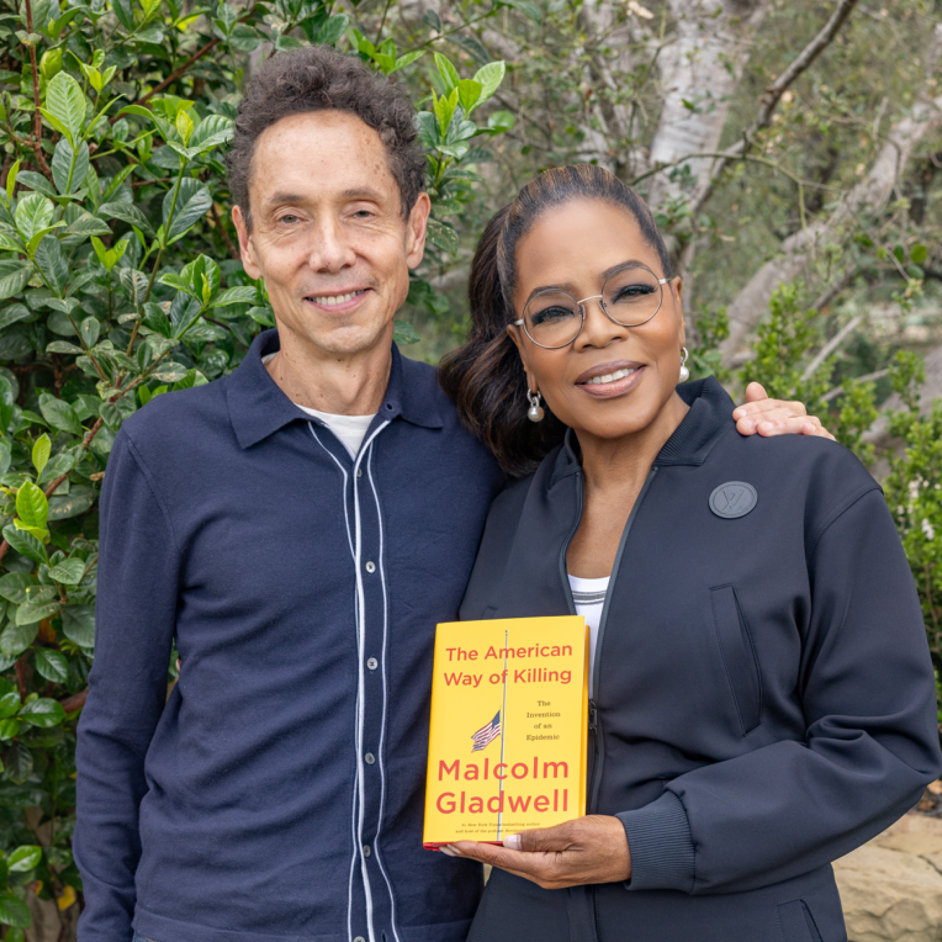 Two people standing outdoors in front of green foliage, one man with curly hair wearing a blue shirt and a woman with glasses and earrings wearing a dark jacket, holding a yellow book titled 'The American Way of Killing' by Malcolm Gladwell.