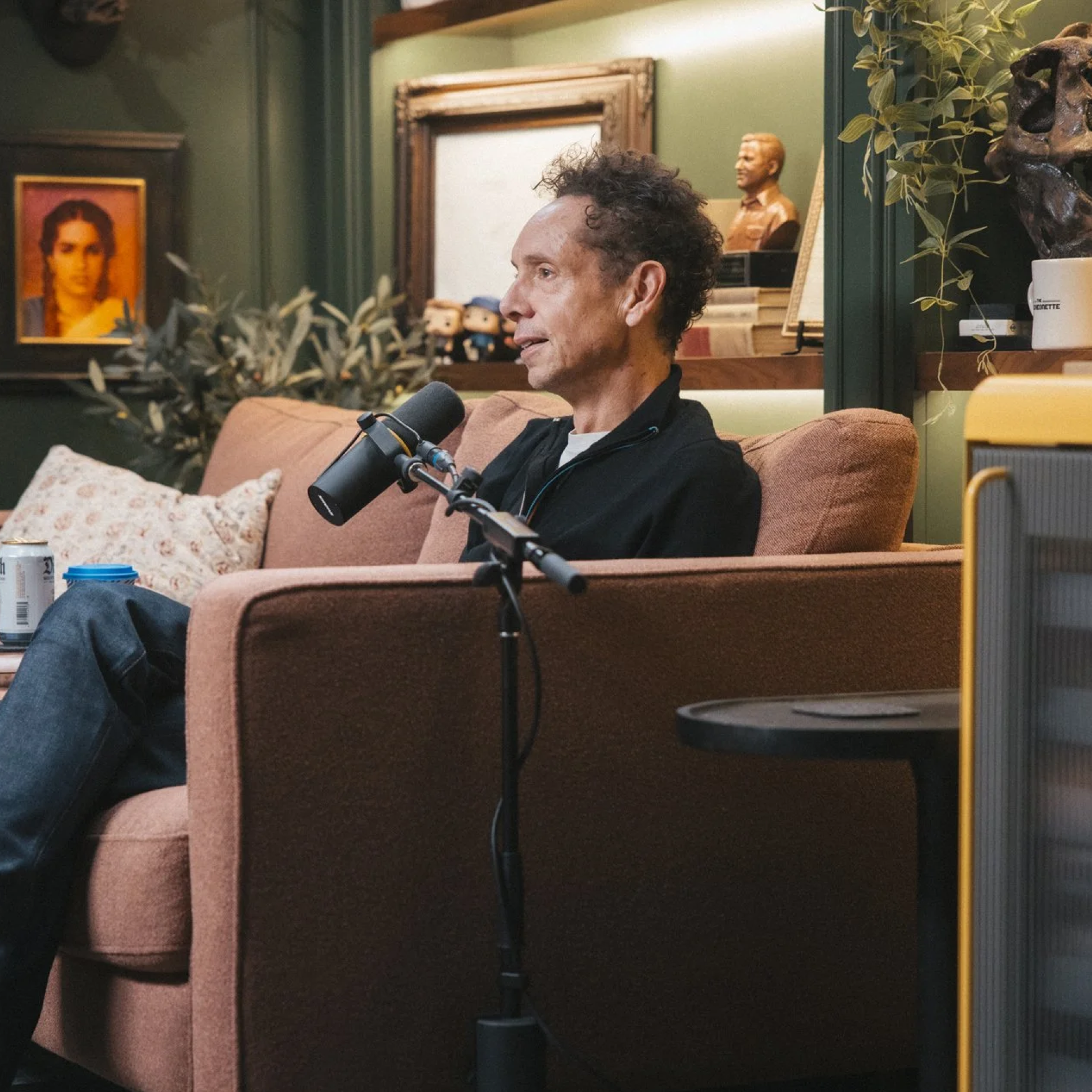 Man with curly hair sitting on a brown couch speaking into a microphone in a cozy, dimly lit room decorated with framed pictures, plants, and small figurines.