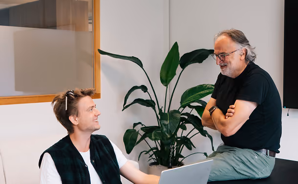 Two men in casual clothes smiling and talking indoors beside a large green plant and laptop.