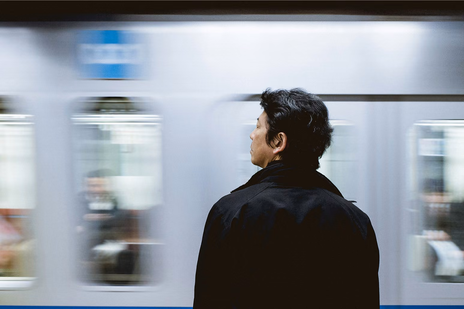 Man with black hair and jacket standing still as a subway train passes by in the background.
