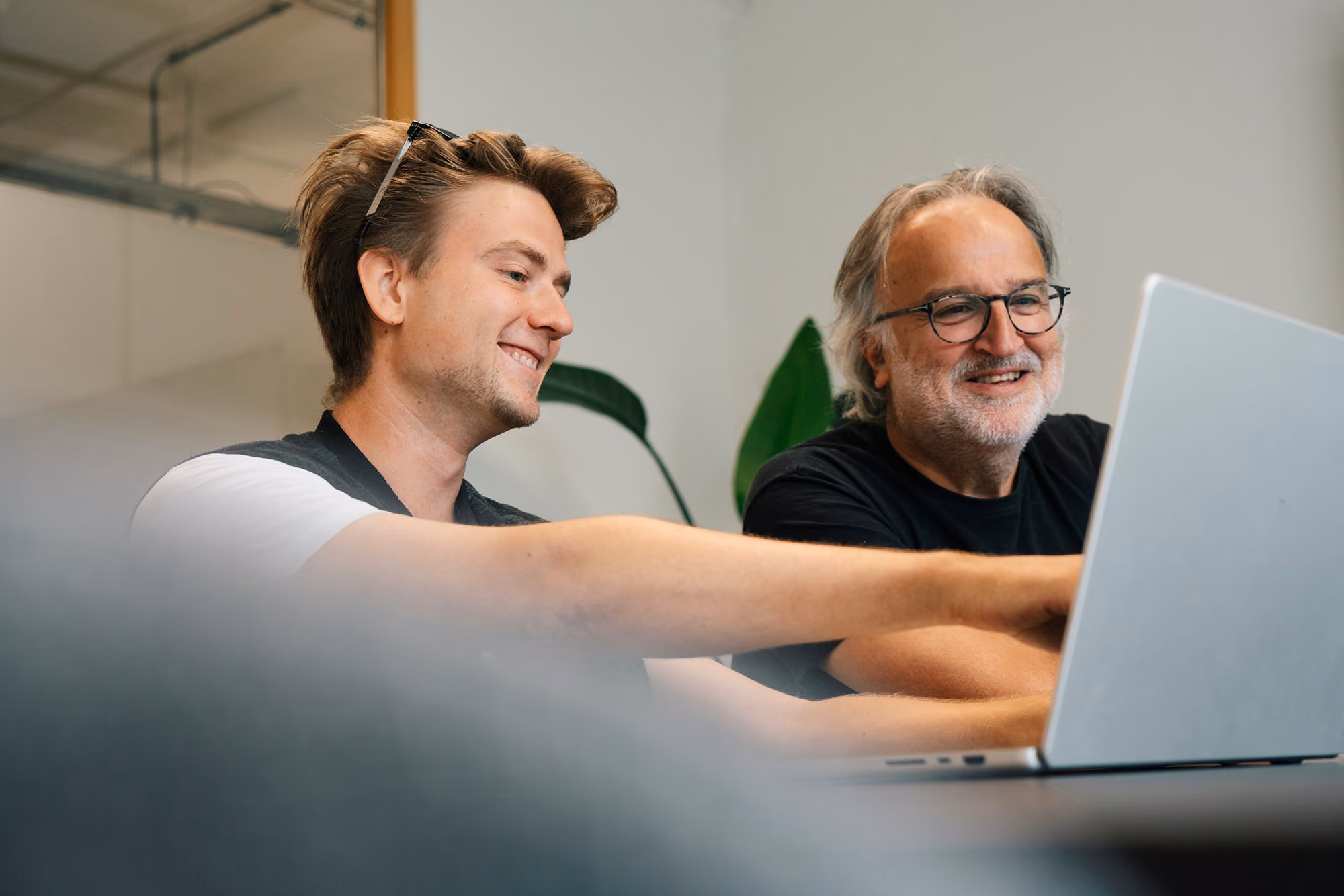 Two men sitting side by side and smiling while looking at a laptop screen in a modern indoor setting.