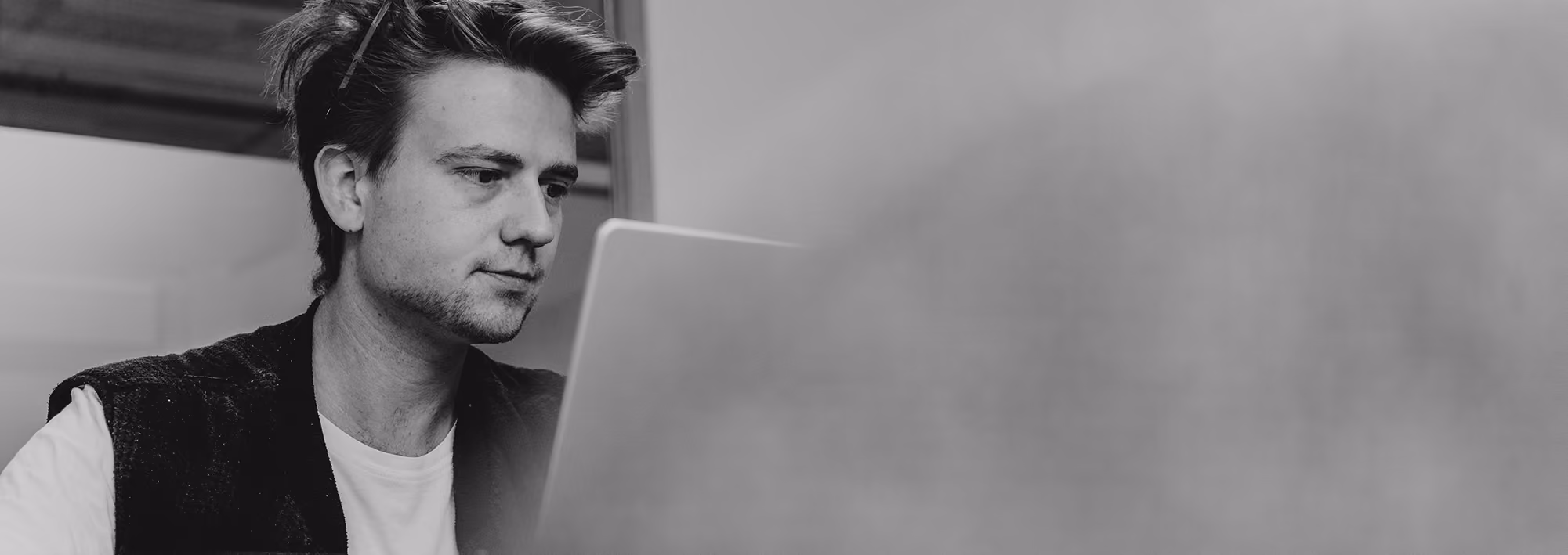 Black-and-white photo of a young man focused on his laptop screen while sitting indoors.