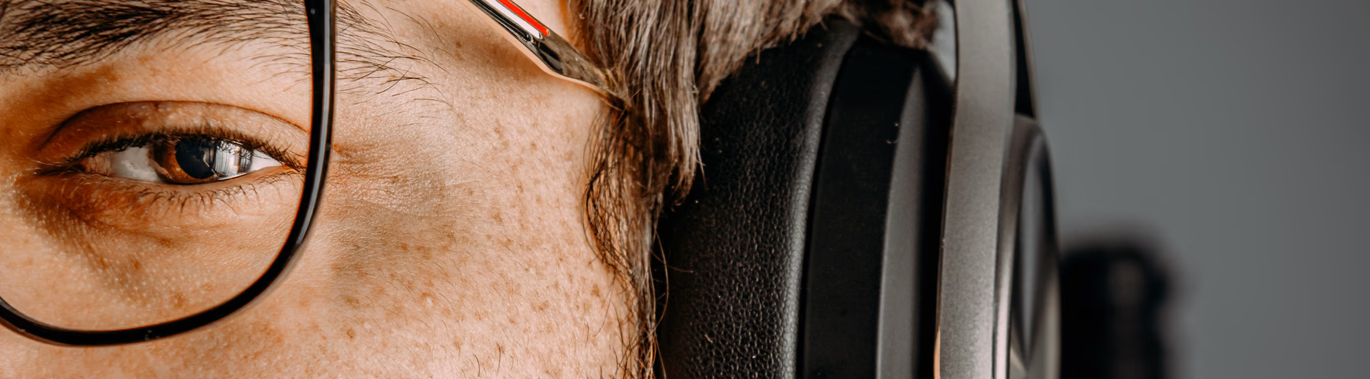 Close-up of a person's eye with glasses and headphones, showing detailed skin texture and freckles.