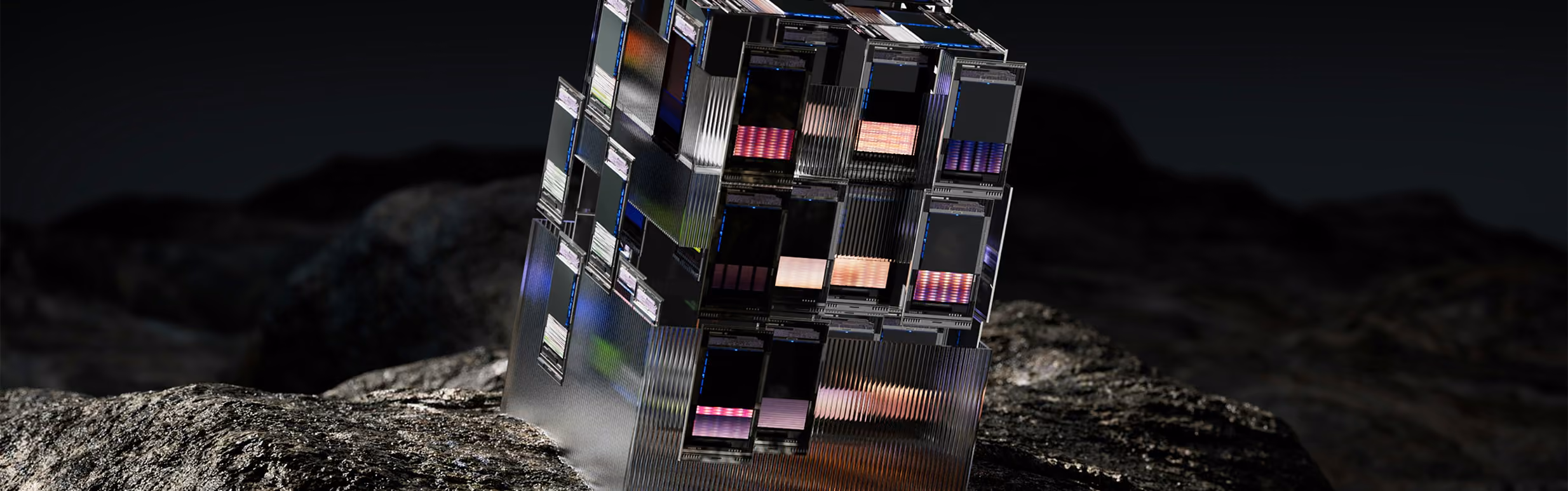 A transparent, cubic structure composed of stacked smaller compartments filled with colorful light elements, positioned on a rough dark rock surface against a black background.