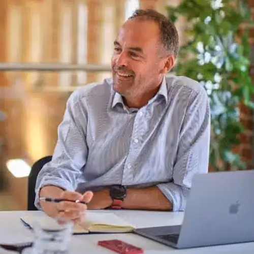 Man, Joe F., in a light shirt, sitting at a desk with a laptop, smiling and holding a pen, with plants in the background.