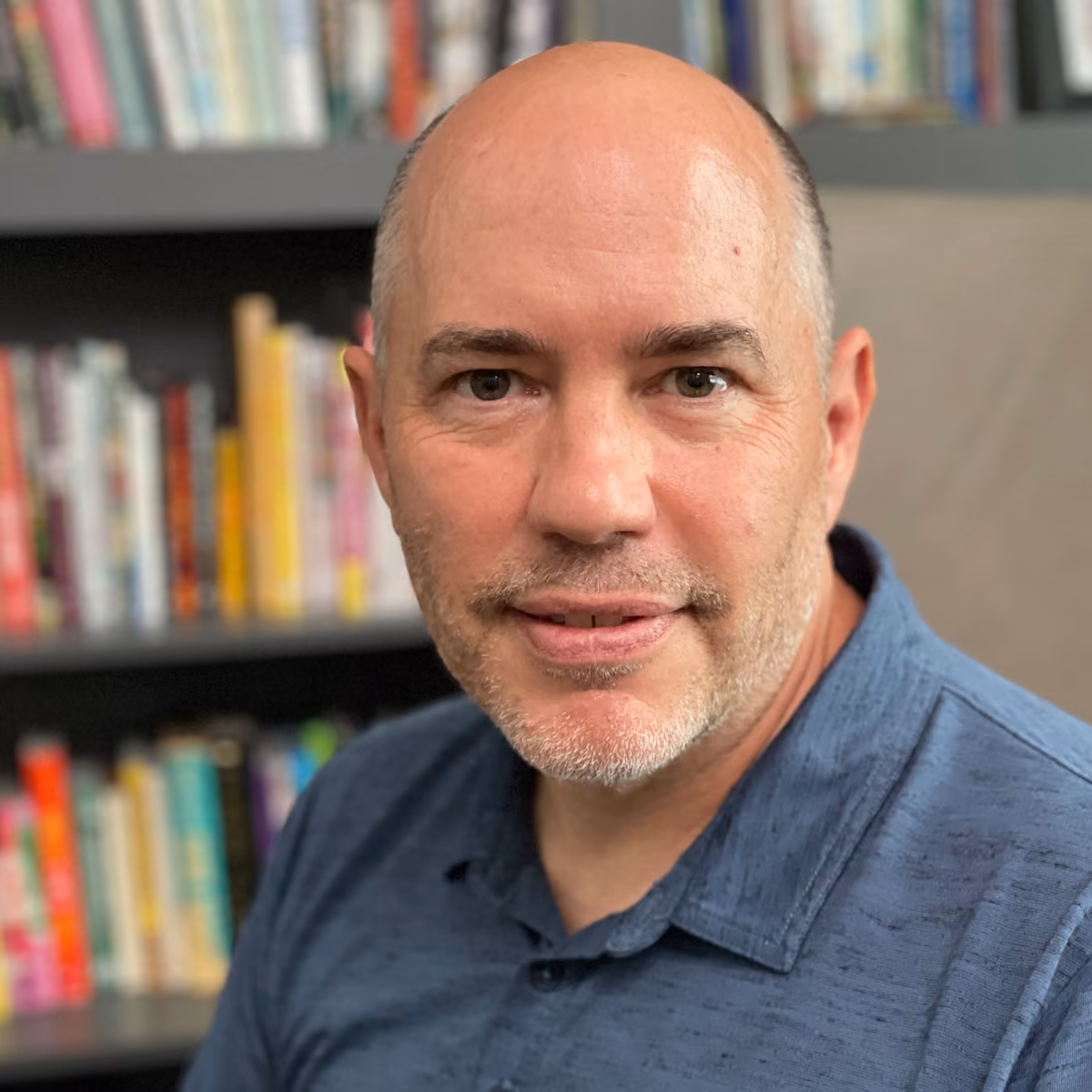 Aleks Szymanski, a middle-aged man with a shaved head and stubble, wearing a blue shirt in front of a bookshelf.