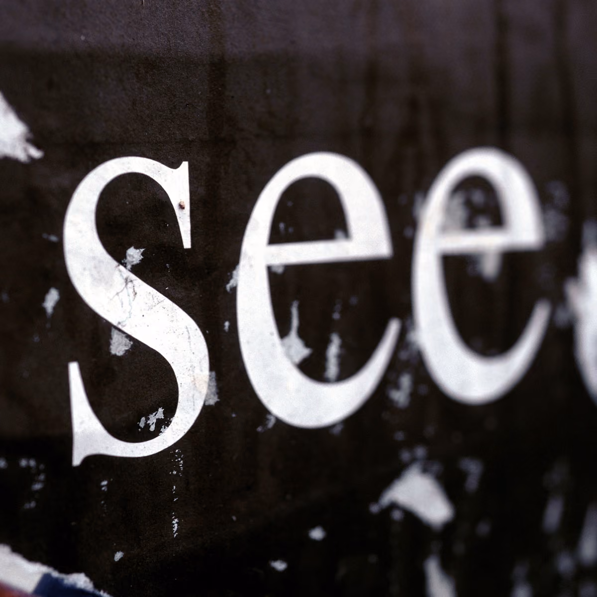 Close-up of white capital letters spelling 'See' on a worn black surface with chipped paint.