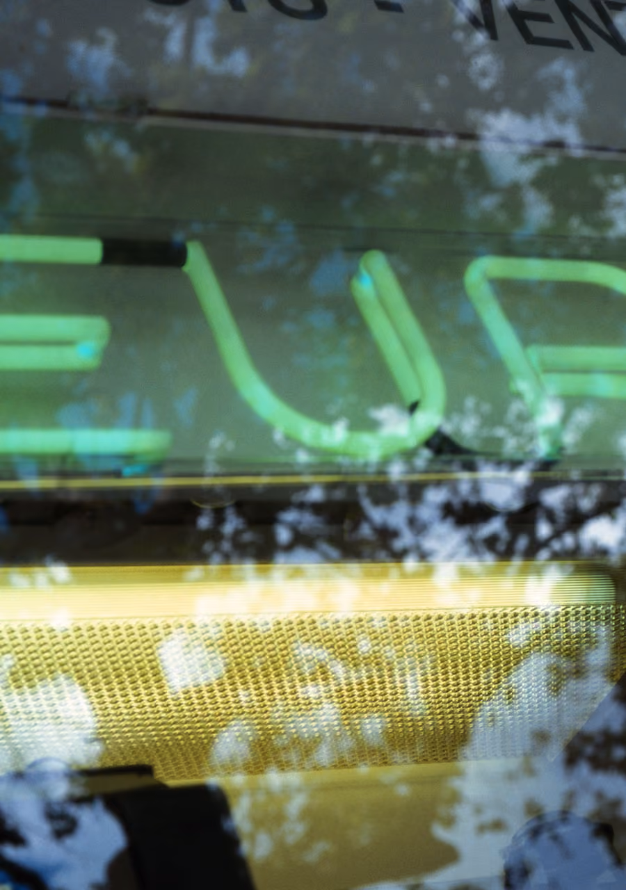Reflection of tree branches over a green neon sign with partially visible letters and a textured yellow surface.