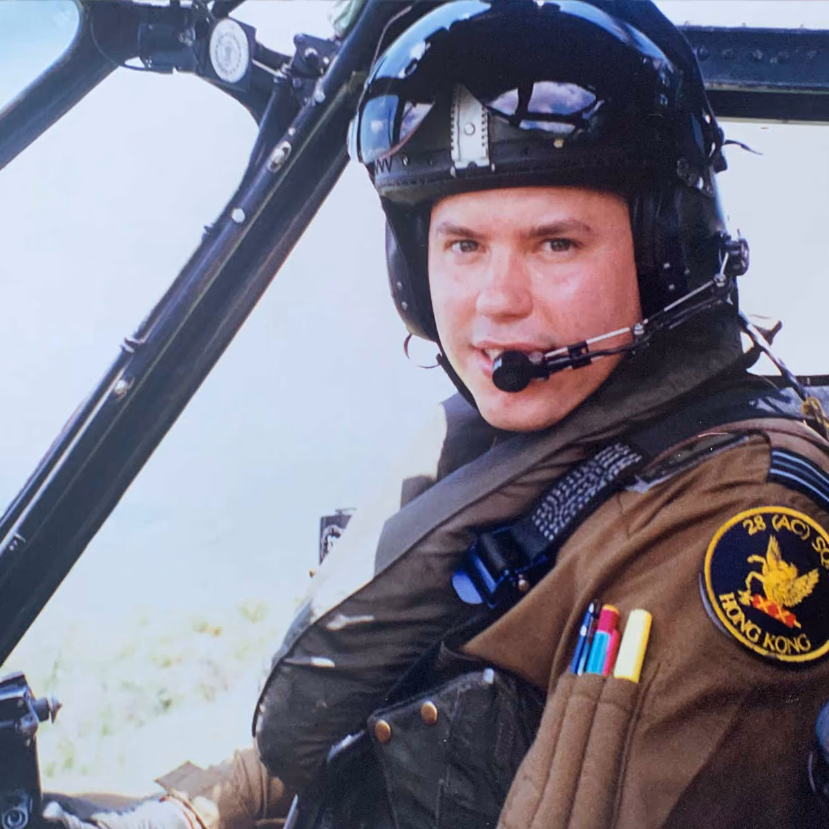 Pilot, Aleks Szymanski, inside cockpit wearing a helmet and headset with a Hong Kong 28 Squadron patch on uniform sleeve.
