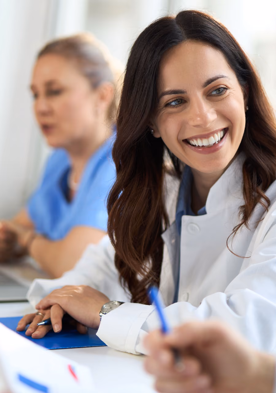 Smiling female doctor in white coat sitting at a table with colleagues in discussion.