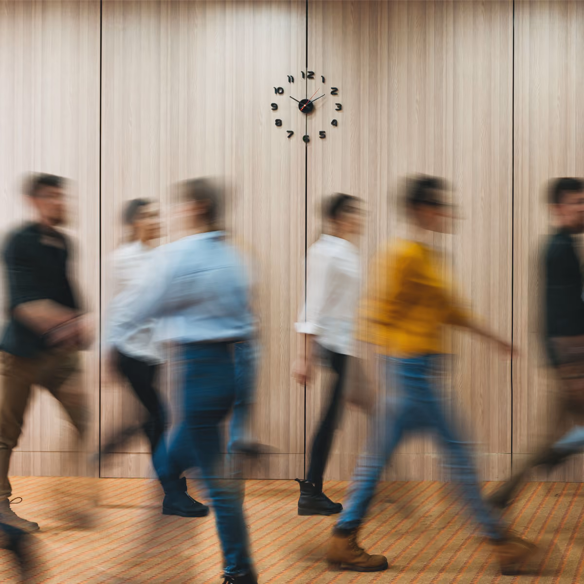 Blurred people walking past a wooden wall clock showing 12:50 inside an indoor space.