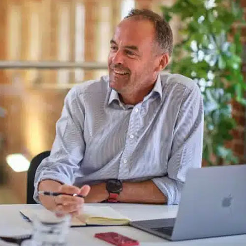 Smiling man in a blue shirt with a laptop and notebook at a desk indoors.