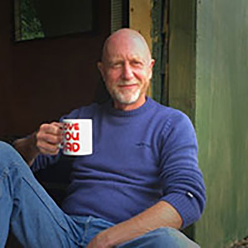 Smiling bald man, Kevin B., in a blue sweater holding a white mug with red text sitting near a green door.