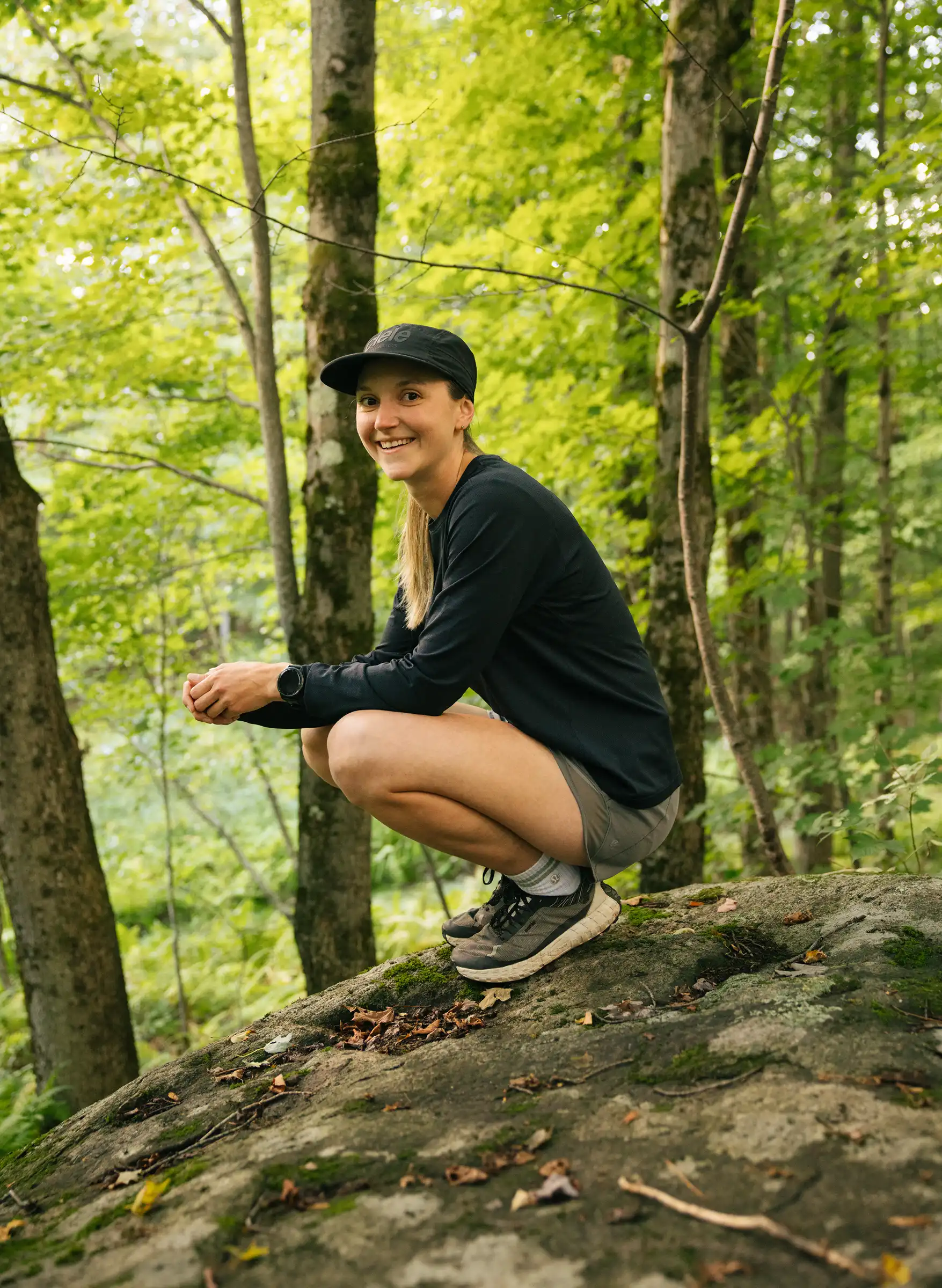 Coureuse accroupie sur un rocher en forêt, souriante, casquette noire