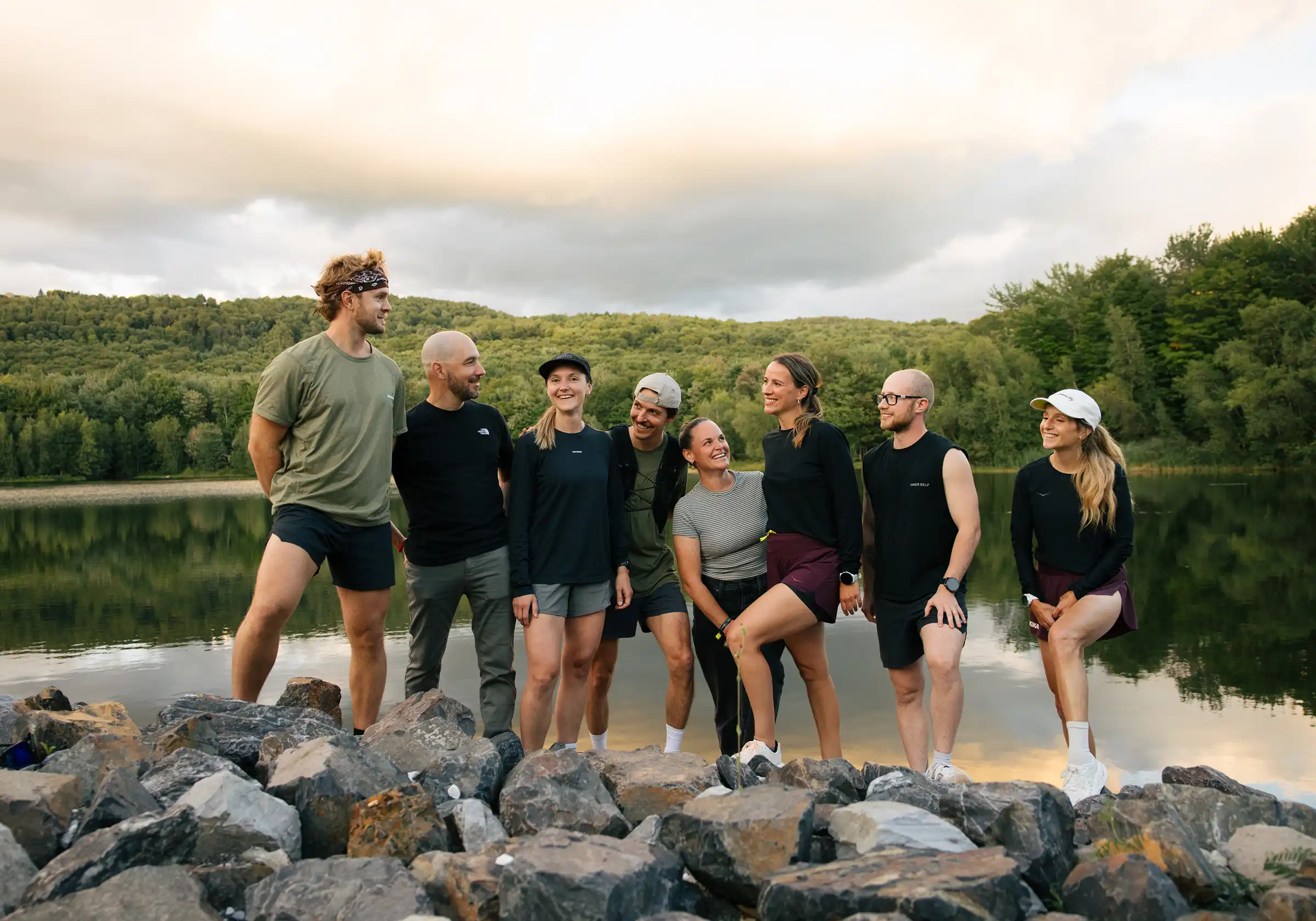 Groupe de huit coureurs réunis sur les rochers au bord d'un lac, forêt et ciel nuageux en arrière-plan