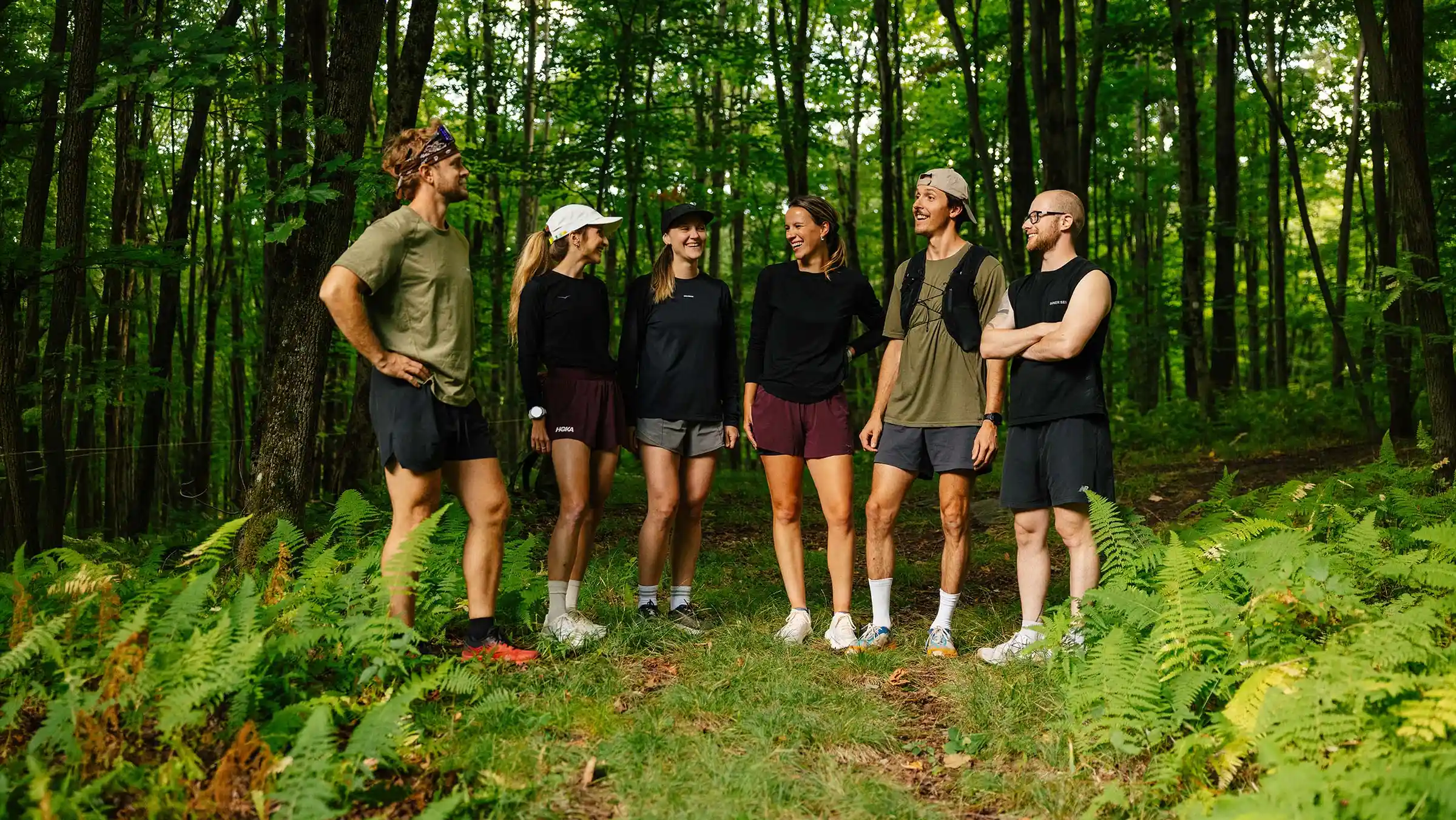 Six coureurs en tenue de trail discutent dans une forêt de feuillus, entourés de fougères